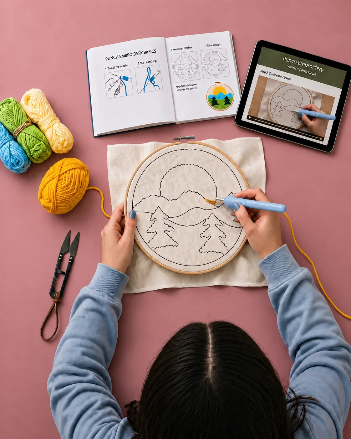 Person using punch needle on embroidery hoop with yarn,instruction book, and tablet with instruction video on pink surface