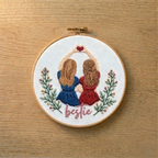 Embroidery hoop with two embroidered figures and the word 'bestie' on a wooden surface