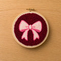 Embroidery hoop with a punch needled pink and white bow design on a red background, placed on a wooden surface.