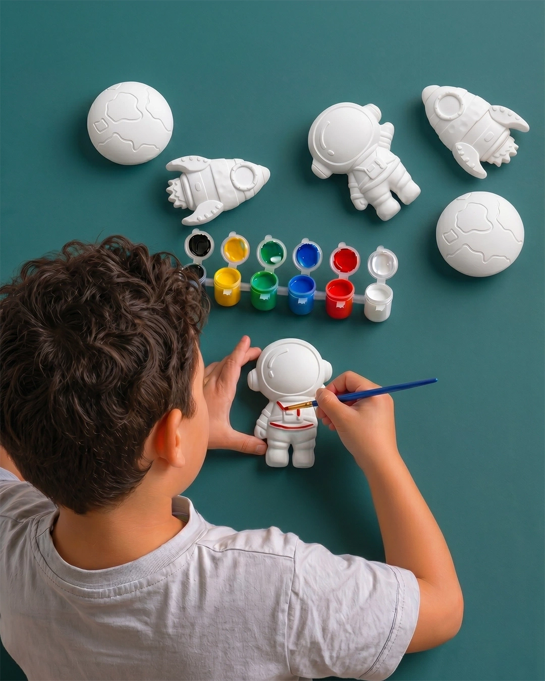 Child painting concrete astronaut figures on a teal surface with paint supplies.