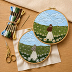 Two wooden embroidery hoop displayed on a rustic wooden surface, showcasing a completed embroidery of a girl in a white dress standing in a daisy field under a blue sky with clouds and other hoop with un-stitched design and threads ,needle, scissors on the wooden surface 