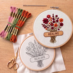 Embroidery hoops with floral designs and 'HEALING' text on a wooden surface with embroidery threads and scissors.