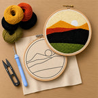 Embroidery hoop with a landscape design, punch needle, thread cutter and yarn balls on a wooden surface