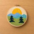 Embroidery hoop with a punch needled landscape design featuring trees, mountains, and a sun on a wooden surface.