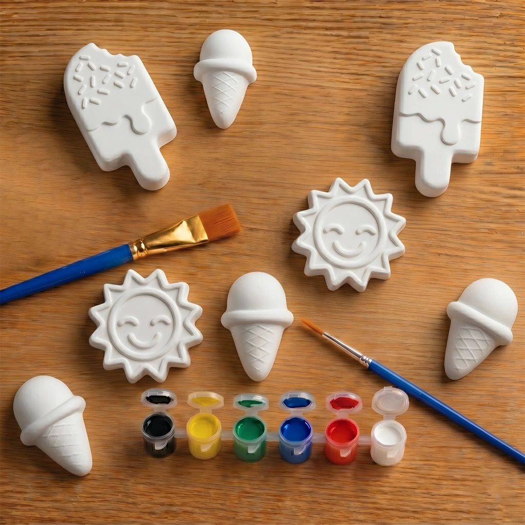 White concrete molds shaped like ice cream cones, suns, and clouds on a wooden surface with paintbrushes and paint.