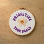 Embroidery hoop with 'Prioritize Your Peace' text and a smiley flower on a wooden surface