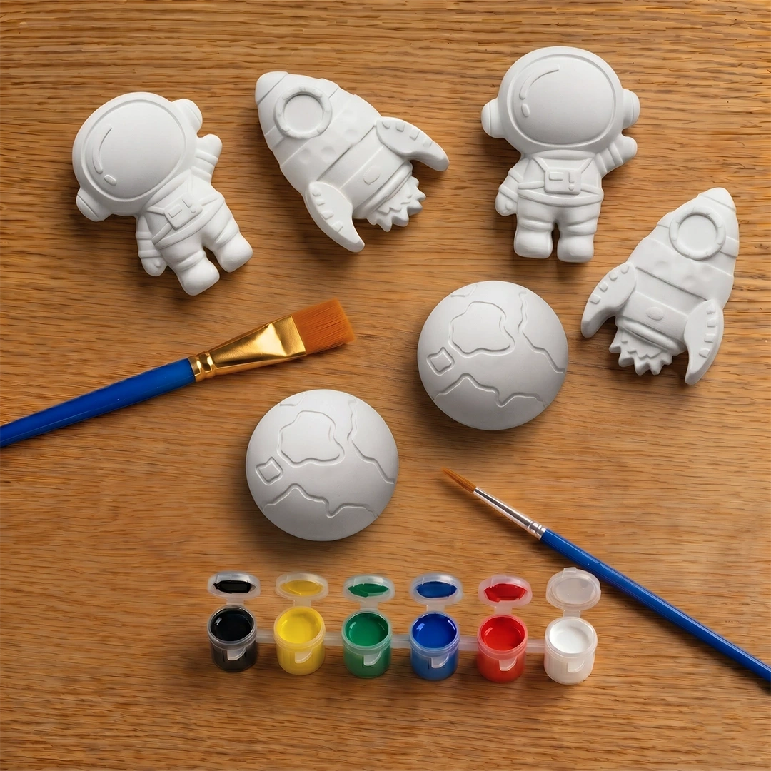 White concrete molds of an astronaut and a rocket on a wooden surface with paintbrushes and paint.