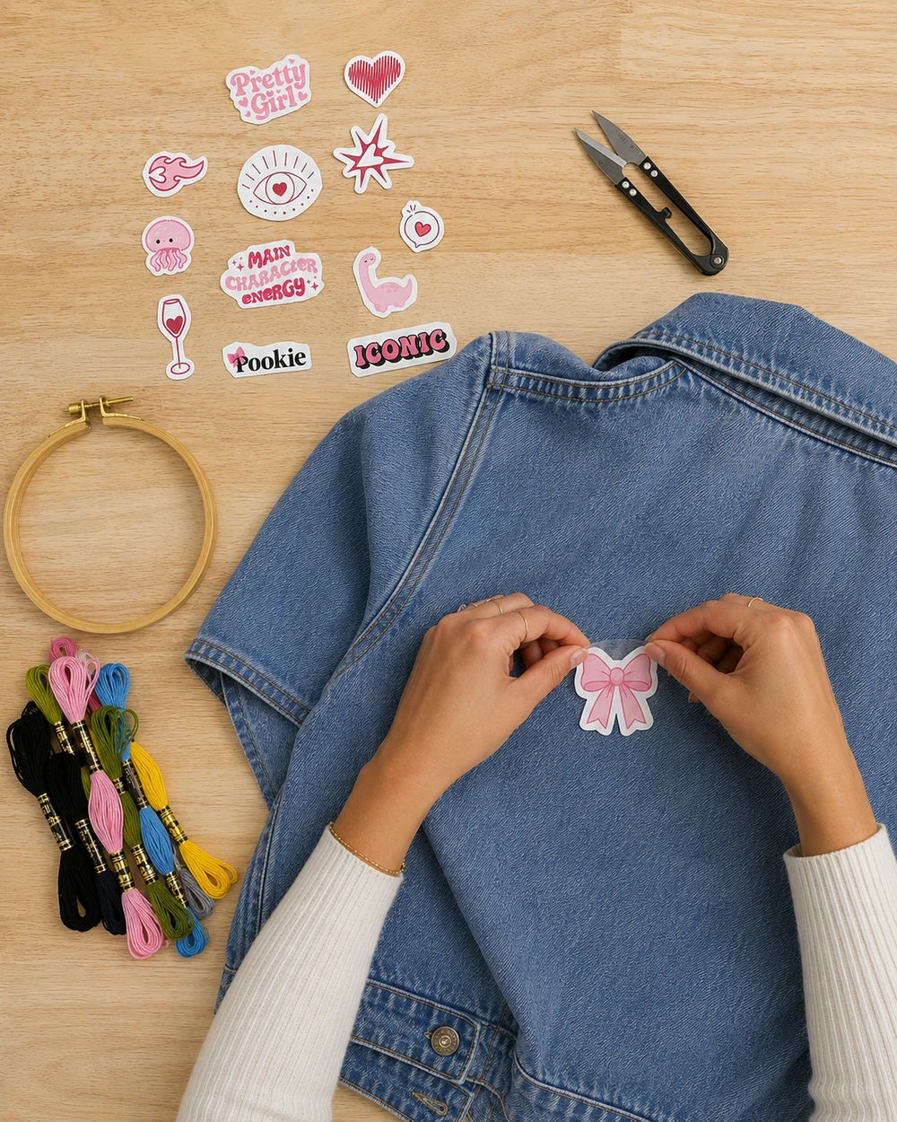 A person's hands are shown applying a pink and white bow stick and stitch onto a blue denim jacket. The scene is styled as a flat-lay on a light wood background, featuring other crafting supplies including an embroidery hoop, spools of thread, and aesthetic themed stick and stitch stickers.