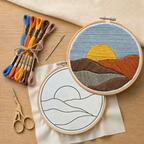 Embroidery hoop with a completed landscape design on a wooden surface with embroidery threads and tools.