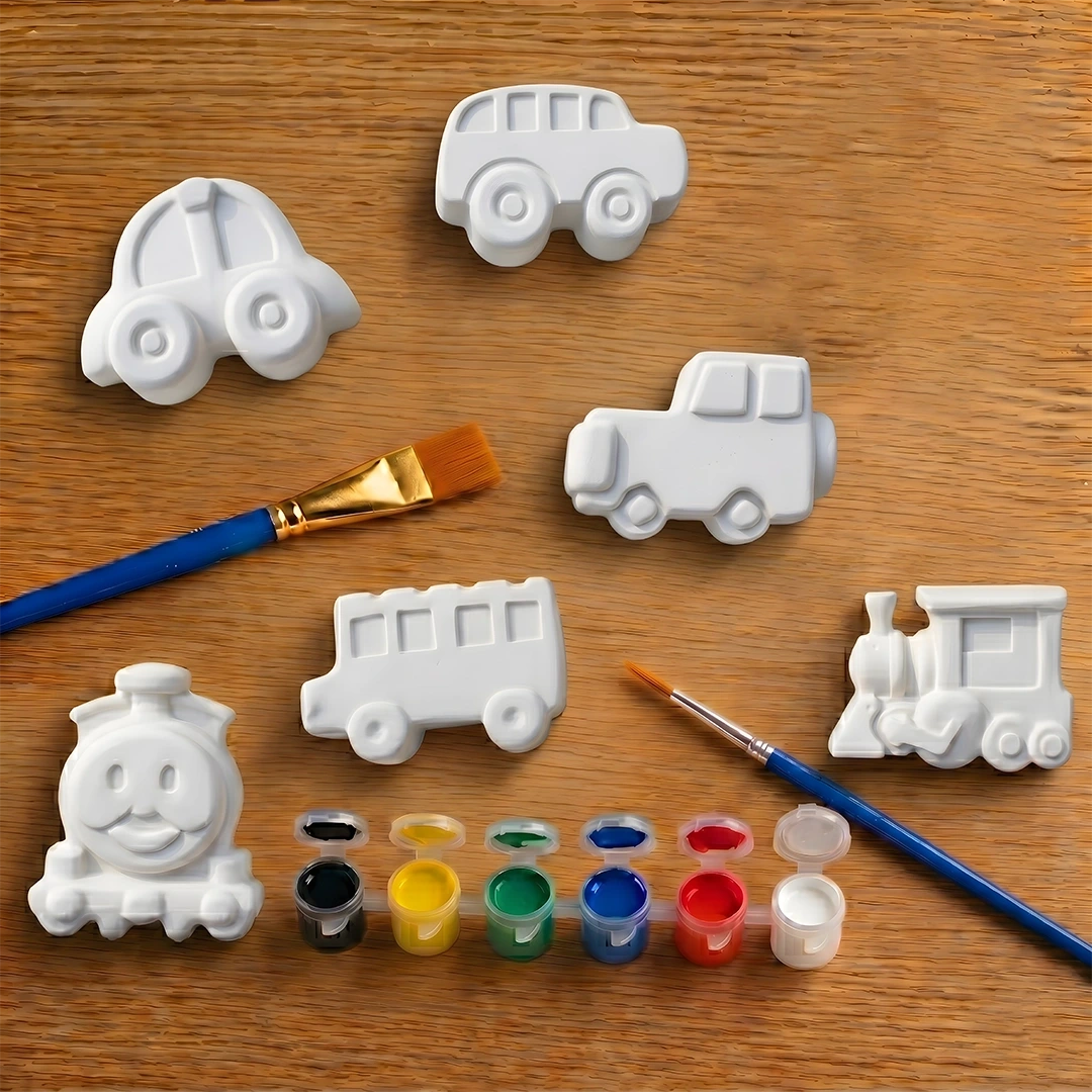 White concrete car, truck, and train molds on a wooden surface with paintbrushes and paint.